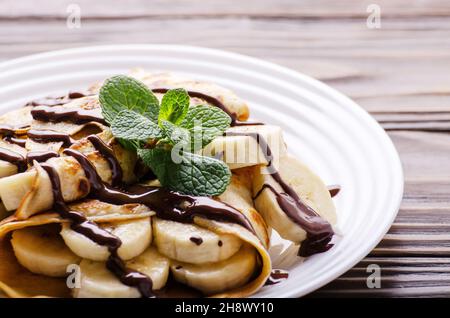 French crepes with chocolate sauce and banana in ceramic dish on wooden kitchen table Stock Photo