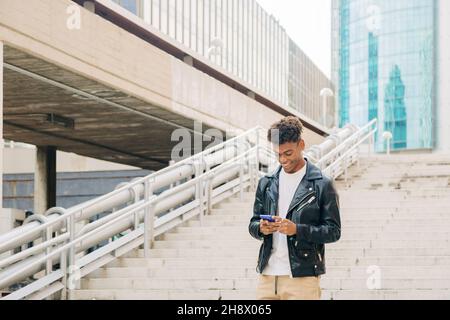 Young African American male standing on stairs of bridge against modern building and browsing mobile phone with smile Stock Photo