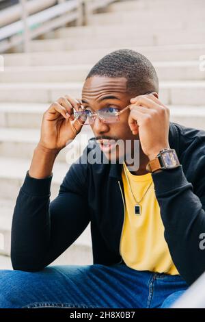 Serious black guy sitting on white street staircase and adjust eyeglasses while looking away Stock Photo
