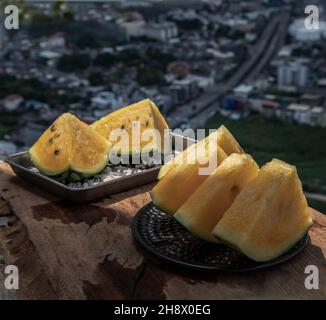 Fresh juicy yellow watermelon cut into triangles on old wooden table at balcony with city view. Tropical fruits. Select focus. Stock Photo