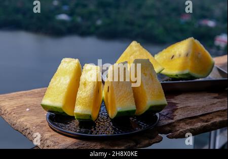 Fresh juicy yellow watermelon cut into triangles on old wooden table at balcony with nature view. Tropical fruits. Select focus. Stock Photo