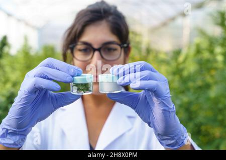 Young female specialist in white robe and latex gloves demonstrating glass bottles with cannabis drugs and plants while working in greenhouse in pharm
