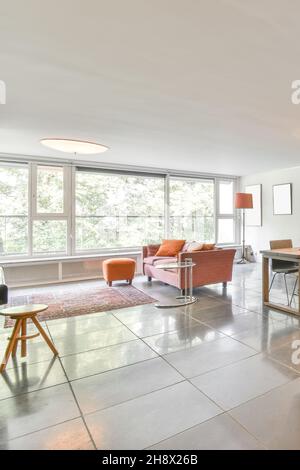 Comfortable sofa with cushions placed on tiled floor near orange pouf in light spacious living room with windows in apartment Comfortable sofa with cushions placed on tiled floor near orange pouf in light spacious living room with windows in apartment