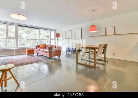 Comfortable sofa with cushions placed on tiled floor near orange pouf in light spacious living room with windows in apartment