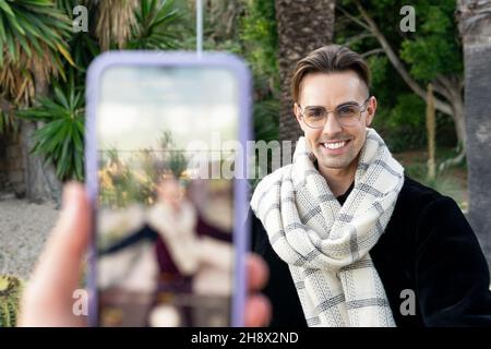 Crop anonymous person taking photo of positive confident young male in trendy scarf and eyeglasses smiling against tropical trees in garden Stock Photo