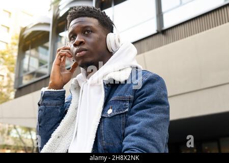 From below of young African American guy with curly hair in stylish denim clothes and white sneakers sitting with headphones listening to music lookin Stock Photo