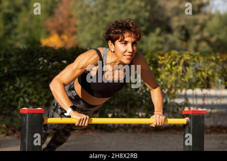 Determined adult ethnic female athlete with short curly hair in sportswear doing push ups on metal bar during outdoor workout in green park on sunny d Stock Photo