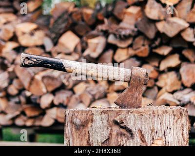 Massive axe in timber stump on background of stacked chopped firewood in countryside yard Stock Photo