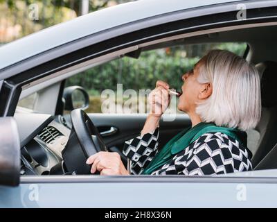 Side view elderly Woman in elegant wear applying lipstick looking in rear view mirror while sitting on driver seat of modern car Stock Photo