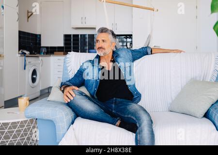 Bearded dreamy old male in denim outfit sitting on couch while looking away thoughtfully against kitchen interior Stock Photo
