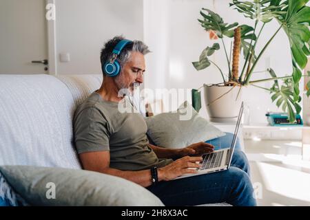Side view of elderly male in headphones sitting on sofa with laptop in modern loft apartment and listening to music Stock Photo