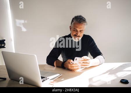 Gray haired male sitting at table with laptop and focusing while browsing smartphone in sunlight Stock Photo