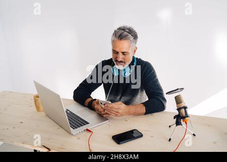 Gray haired male broadcaster sitting at table with laptop and focusing while browsing smartphone in sunlight Stock Photo