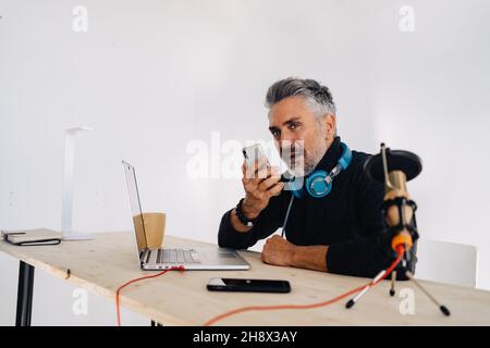 Old stylish male broadcaster with beard sitting at table with laptop and looking away while recording audio message on mobile phone Stock Photo