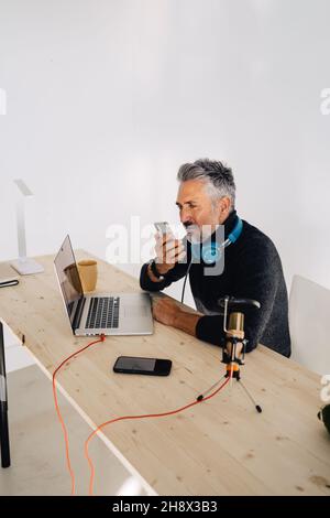 Old stylish male broadcaster with beard sitting at table with laptop and looking away while recording audio message on mobile phone Stock Photo