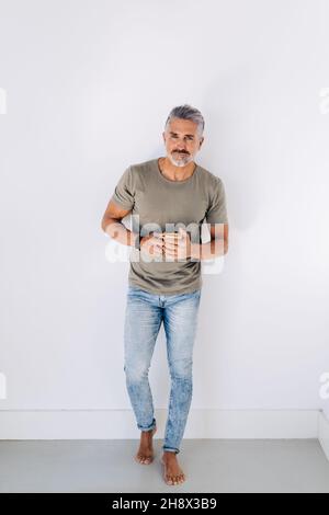Thoughtful barefoot aged male in casual clothes leaning on wall looking away while drinking hot beverage Stock Photo