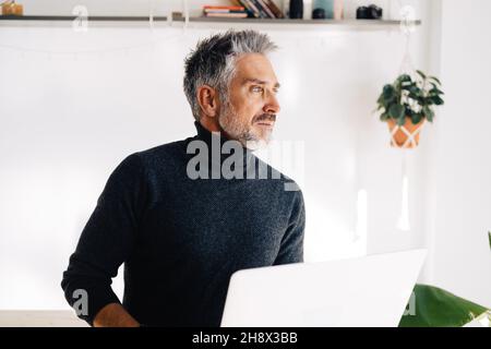 Senior bearded broadcaster standing near desk and using laptop in hands while working on project in home studio Stock Photo