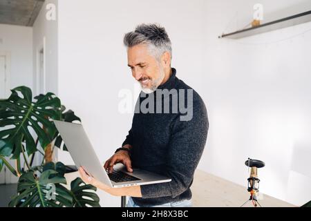 Cheerful senior bearded broadcaster standing near desk and using laptop in hands while working on project in home studio Stock Photo