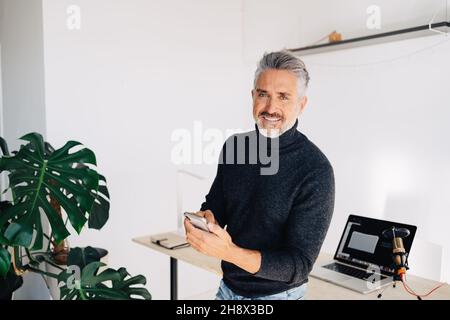 Positive gray haired male standing against table with laptop and microphone and using mobile phone while preparing for recording podcast Stock Photo