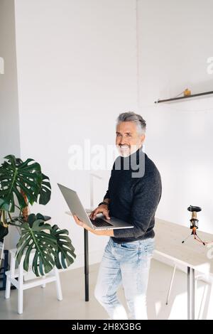 Cheerful senior bearded broadcaster standing near desk and using laptop in hands while working on project in home studio looking at camera Stock Photo