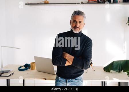 Content elderly broadcaster standing near table with laptop and headphones and looking at camera with crossed arms in light room Stock Photo