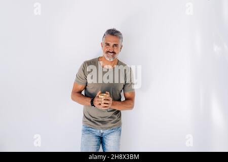 Smiling aged male in casual clothes leaning on wall looking away while drinking hot beverage Stock Photo