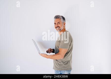 Elderly bearded smiling male looking at camera attentively while standing against white wall and browsing laptop in hand in light room Stock Photo