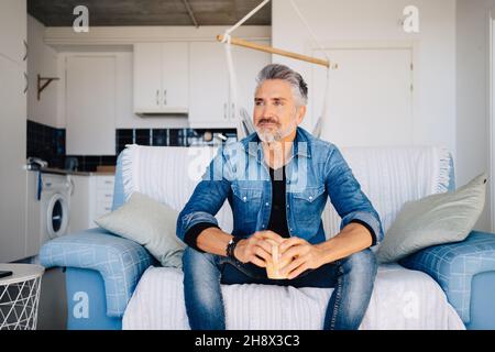 Bearded dreamy old male in denim outfit sitting on couch while drinking tea and looking away thoughtfully against kitchen interior Stock Photo