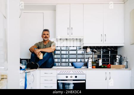 Elderly thoughtful man holding cup of tea while sitting on counter in kitchen with white cabinets and looking away Stock Photo