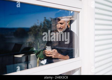 Through glass of old bearded man looking out window and browsing smartphone while working in office in sunny day Stock Photo