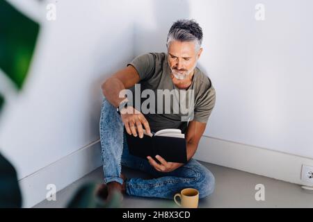 High angle of concentrated aged man in casual clothes drinking tea and reading book while sitting on floor in corner Stock Photo