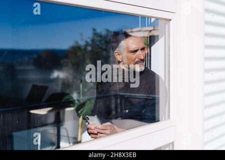 Through glass of old bearded man looking out window and browsing smartphone while working in office in sunny day Stock Photo