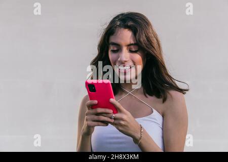 Isolated happy Hispanic female smiling and using red cellphone while texting against white backdrop Stock Photo