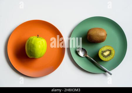 From above of fresh tasty green apple and ripe whole and halved kiwi placed on two colored ceramic plates with silver spoon on white table Stock Photo