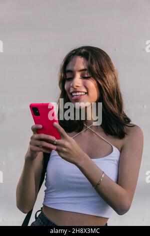 Happy Hispanic female smiling and using red cellphone while texting against white backdrop Stock Photo
