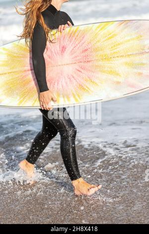 Crop unrecognizable female surfer in wetsuit standing on wet seashore with surfboard near waving sea in coastal area on summer day Stock Photo