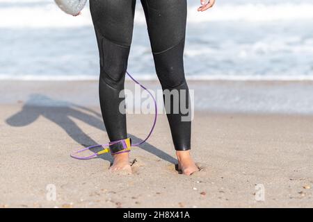 Crop unrecognizable female surfer in wetsuit standing on wet seashore with surfboard near waving sea in coastal area on summer day Stock Photo