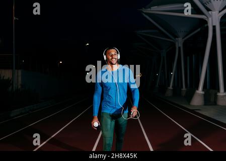 African athlete listening music and walking towards the camera on the running track at night Stock Photo