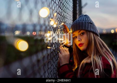 Pretty blonde young Woman with wool hat leaning on the fence and holding a light garland of lights looking at the camera Stock Photo