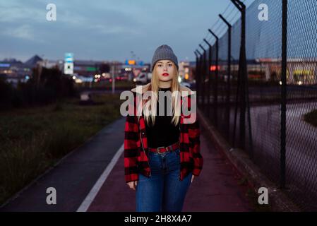 Pretty blonde young Woman with wool hat and plaid jacket standing on the street in the evening Stock Photo