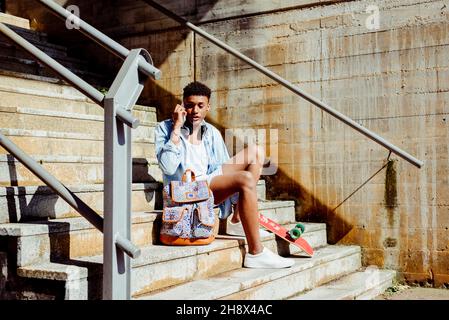 Young ethnic black man sitting on stairs on phone call on smartphone sitting on the street Stock Photo