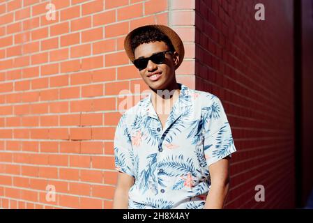 Trendy young black man with sunglasses and a hat posing on the street next to a brick wall looking at camera Stock Photo