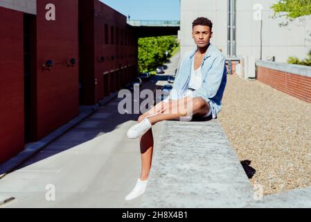 Young ethnic man sitting on the roof cornice of a building looking at camera Stock Photo