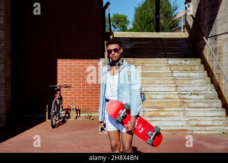 Young ethnic black man with sunglasses standing on the street holding a skateboard and mobile phone Stock Photo