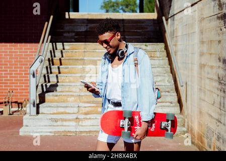 Young ethnic black man with sunglasses browsing on smartphone on the street and holding a skateboard Stock Photo