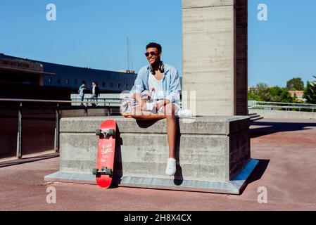 Young black man with sunglasses sitting on stone bench in the street using the phone with headphones on neck Stock Photo