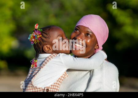 Adorable little black girl with Afro braids hugging and kissing cheek of happy mother in casual clothes and bandana standing in nature Stock Photo