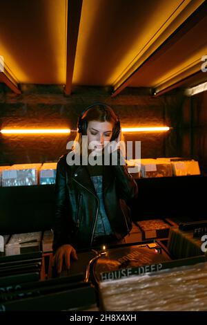Front view of young female listening to music in headphones while standing near racks with collection of vinyl records in store with glowing lights Stock Photo