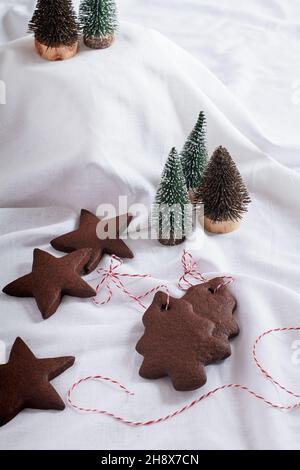 Christmas tree and star shaped chocolate gingerbread biscuits (cookies) on white material with miniature christmas trees. Stock Photo