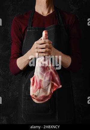 Butcher holds raw lamb leg in hands over dark background Stock Photo